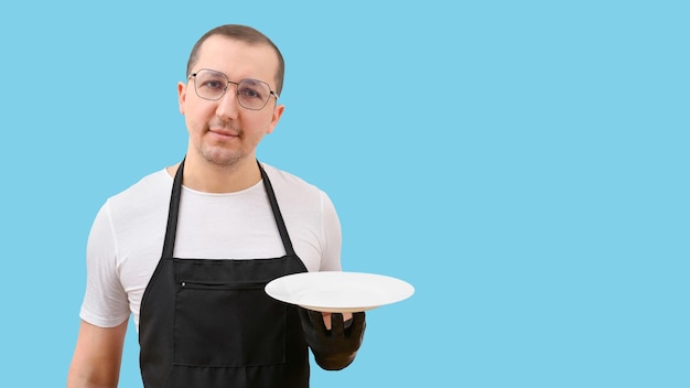 Portrait of a waiter in a black apron holding an empty plate on a blue background