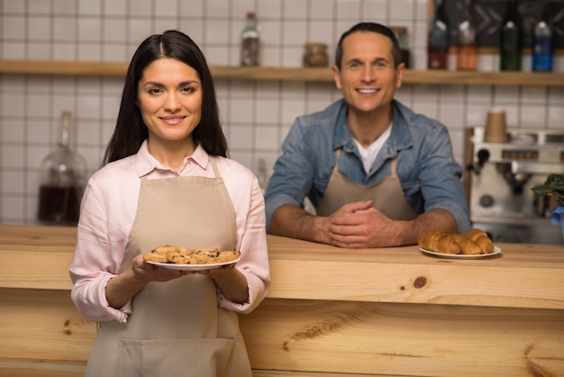 Portrait of waitress holding cookies on the plate and looking at camera with waiter on the background