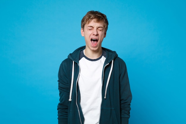 Portrait of weird young man in casual clothes keeping eyes closed screaming isolated on blue wall.