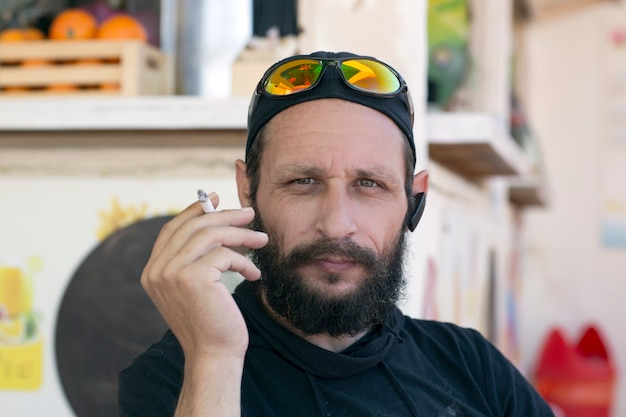 Portrait of a white cool man smoking cigarette wearing sunglasses, cap and hood jacket on the street. Hipster American man smoking in real life. handsome Jewish man bearded man mustache and black hat