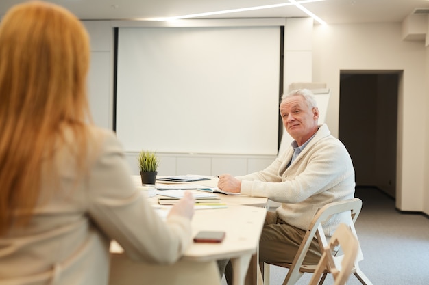 Photo portrait of white-haired senior businessman sitting at table in conference room and listening to female colleague or manager while planning work project, copy space