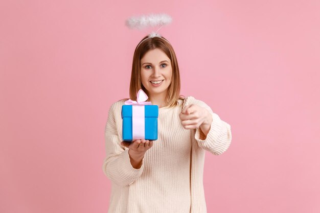 Portrait of winsome young blond woman with halo over head holding blue gift box in hands and pointing at camera wearing white sweater Indoor studio shot isolated on pink background