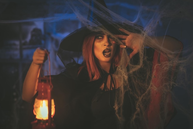 Portrait of witch with awfully face and lighted lantern in her hand in creepy foggy surroundings sends evil.
