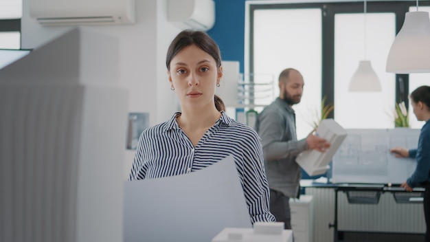 Portrait of woman architect studying blueprints plans on paper to design building model for urban development. Architectural engineer looking at construction layout sketch and maquette.