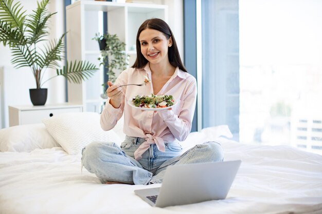 Portrait of woman on bad at home and using laptop while eating healthy salad