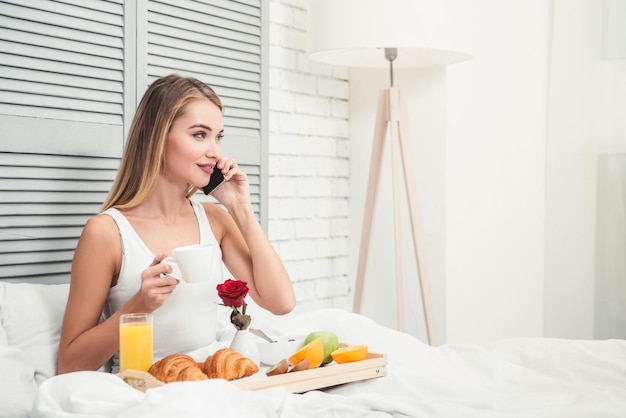 Ritratto di donna nel letto che parla sullo smartphone mentre fa colazione