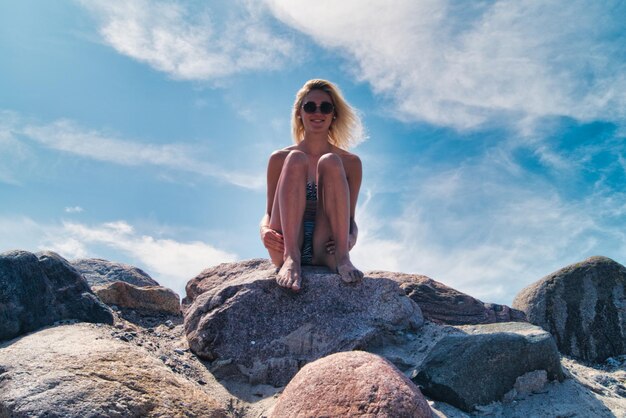 portrait of woman in bikini wearing sunglasses while sitting on rock against sky