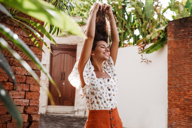 Portrait of woman in bright skirt and blouse with floral print raising her hair Girl in high spirits poses against background of house with old wooden doors and tropical trees
