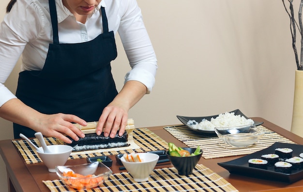 Ritratto di donna chef che arrotola un sushi giapponese con riso, avocado e gamberetti su un foglio di alga nori