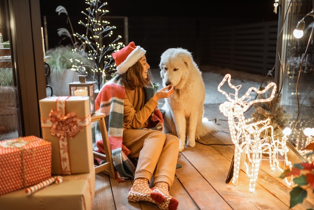 自宅の美しく装飾されたテラスで新年の休暇を祝うかわいい犬と一緒にクリスマス帽子と格子縞の女性の肖像画、ジンジャーブレッドクッキーを犬に与えます