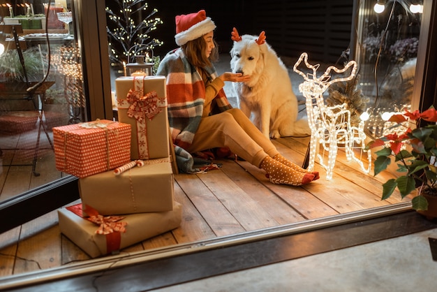 自宅の美しく装飾されたテラスで新年の休暇を祝うかわいい犬と一緒にクリスマス帽子と格子縞の女性の肖像画、ジンジャーブレッドクッキーを犬に与えます