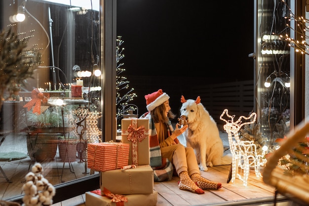 Ritratto di una donna con cappello natalizio e plaid con il suo simpatico cane che celebra le vacanze di capodanno sulla terrazza splendidamente decorata di casa, dando da mangiare al cane con biscotti di pan di zenzero