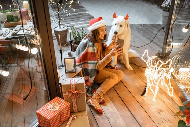 クリスマスの帽子をかぶった女性と、家で年末年始を祝うかわいい犬と格子縞の女性の肖像画、ジンジャーブレッドクッキーを犬に与え、自分撮り写真を作成する