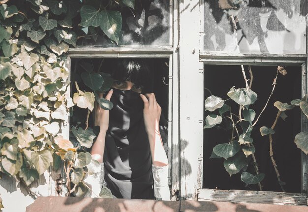 Photo portrait of woman covering face while standing by window amidst plant