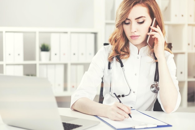 Portrait of a woman doctor with red hair talking on the phone in her office and taking notes in a clipboard. Toned image