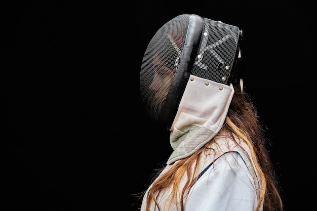 Portrait of woman dressed in white fencing costume practicing with the sword on black background.