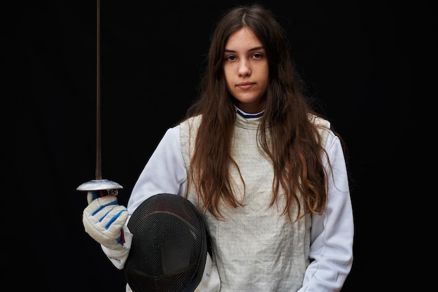 Portrait of woman dressed in white fencing costume practicing\
with the sword on black background.