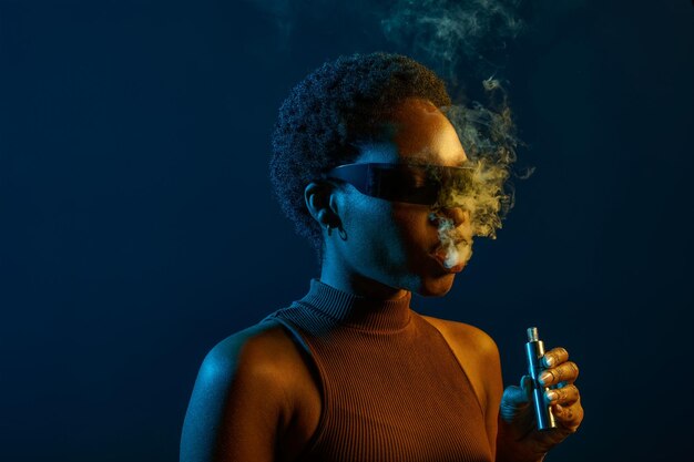 Portrait of woman in eyeglasses smoking vape electronic cigarette over dark studio background