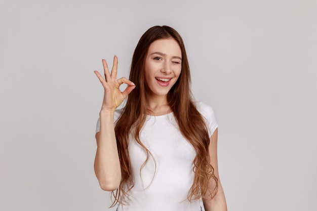 Portrait of woman gesturing okay sign, completely agree with suggestion, confident in success, expressing playful emotions, wearing white T-shirt. Indoor studio shot isolated on gray background.