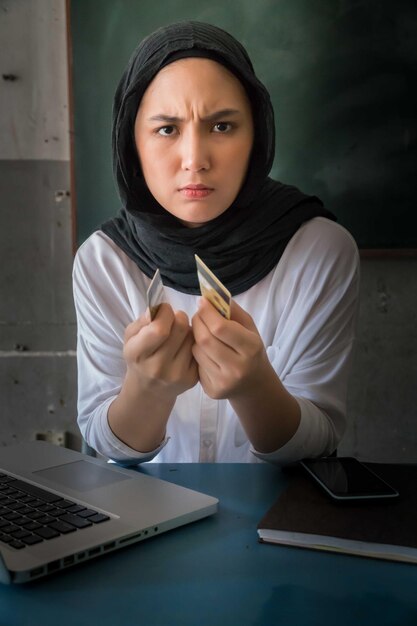 Photo portrait of woman in hijab holding broken credit card at home