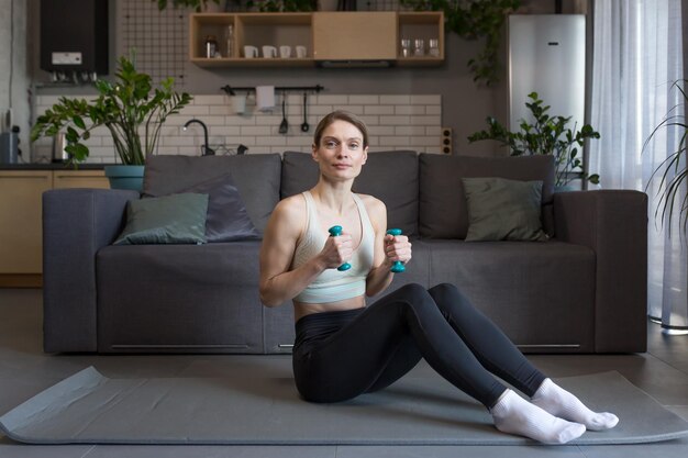 Portrait of woman at home performing morning exercise with dumbbells looking at camera smiling