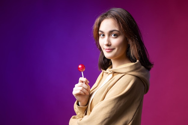 Portrait of a woman in a hoodie girls licking a red round lollipop with beautiful makeup on a purple trendy background in the studio