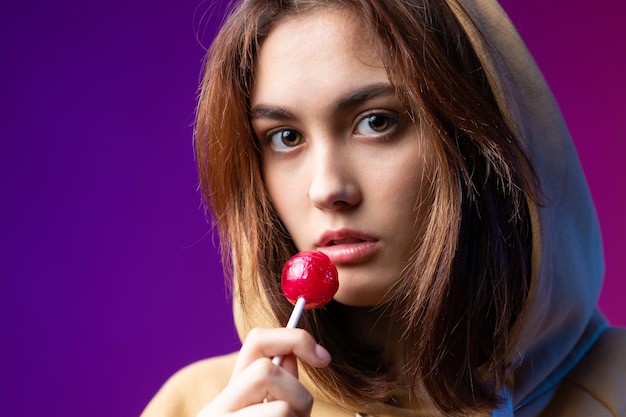 Portrait of a woman in a hoodie girls licking a red round lollipop with beautiful makeup on a purple trendy background in the studio