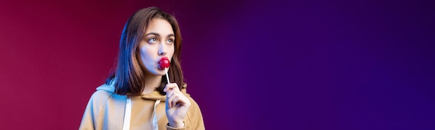 Portrait of a woman in a hoodie girls licking a red round lollipop with beautiful makeup on a purple trendy background in the studio