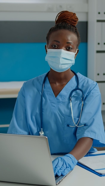 Portrait of woman nurse sitting at desk and using laptop in medical office room. Young adult wearing uniform, face mask and gloves working on healthcare during covid 19 pandemic