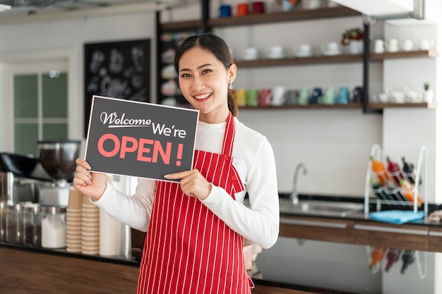 Portrait of woman owner standing at her coffee shop gate with showing open signboard