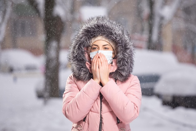 Portrait of woman in pink walking down the street in winter under snowfall in a protective mask to protect against infectious diseases Protection against colds flu air pollution Health concept