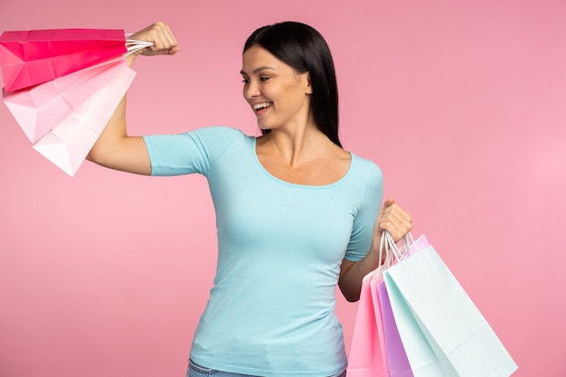 Portrait of woman showing her muscles and holding paper bags, shopping and pleasant prices. Indoor studio shot isolated on pink background