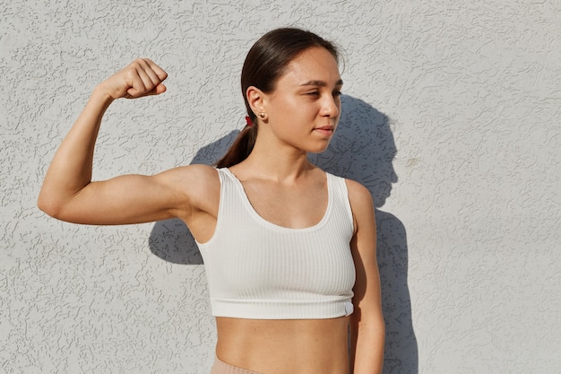 Portrait of woman showing her trained body to the camera, raised arm, shows her hand, biceps, looking away, wearing white top, standing near light wall outdoor.