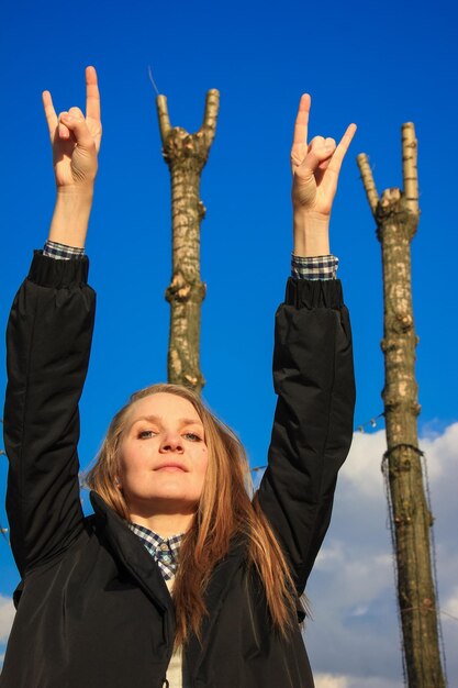 Foto ritratto di una donna che mostra un segno di roccia con le braccia alzate contro il cielo blu