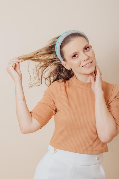 Portrait of woman smiling with hand in hair. studio photo.\
woman wears skin-colored blouse and white pants and two tiaras in\
her hair.