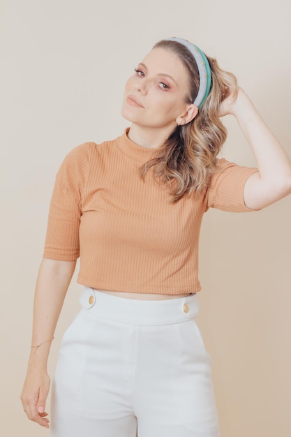 Portrait of woman smiling with hand in hair. Studio photo. Woman wears skin-colored blouse and white pants and two tiaras in her hair.