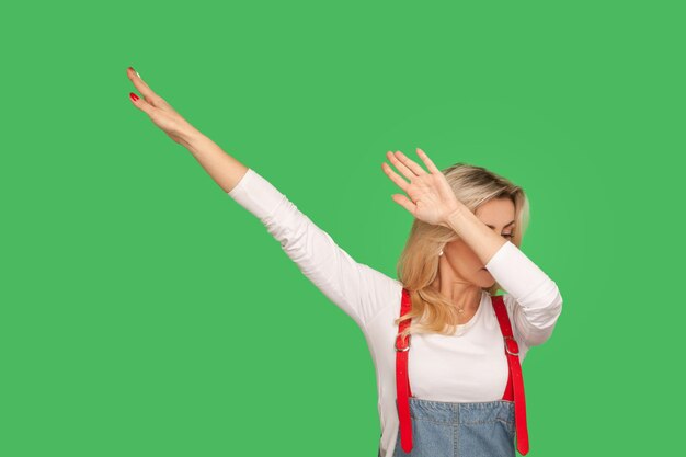 Portrait of woman in stylish denim overalls raising hands in dab dance pose, celebrating success, showing gesture of triumph, famous internet move. indoor studio shot isolated on green background