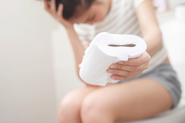 Portrait of a woman suffers from diarrhea his stomach painful ache and problem hand hold tissue paper roll in front of toilet bowl constipation in bathroom Hygiene health care concept