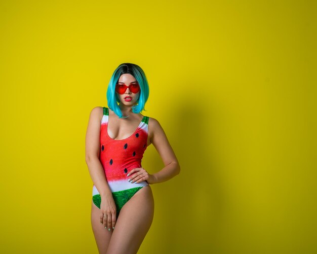Portrait of a woman in a swimsuit with a picture of a watermelon and red sunglasses in the shape of hearts Stylish girl in a colored short wig posing in the studio on a yellow background