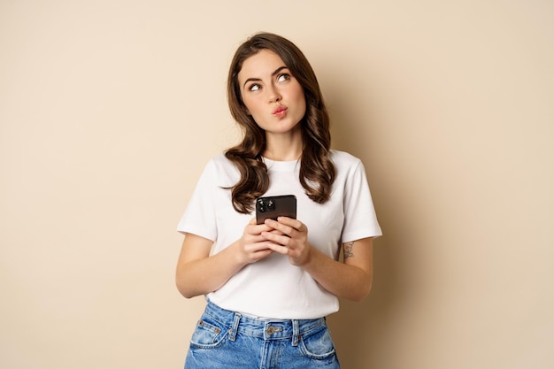 Portrait of woman thinking holding mobile phone and looking up with thoughtful face standing over be...