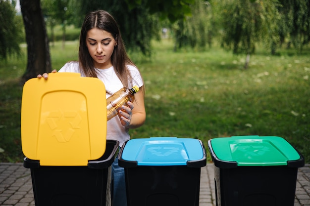 Ritratto di donna che getta una bottiglia d'acqua di plastica vuota in contenitori per il riciclaggio dell'albero del contenitore di riciclaggio all'aperto