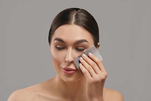 Portrait of woman using facial oil blotting paper on grey background