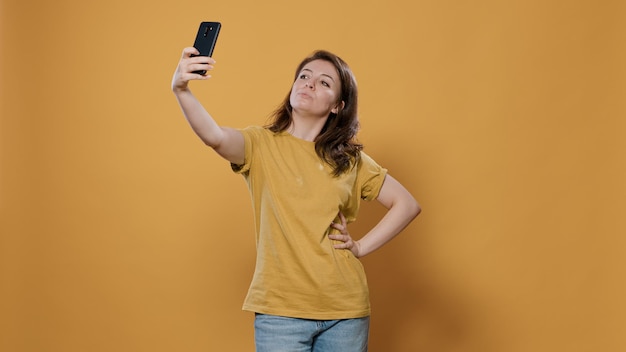 Portrait of woman using smartphone to take a selfie feeling confident and beautiful striking multiple poses in studio. Person using mobile phone taking picture using camera to post on social media.