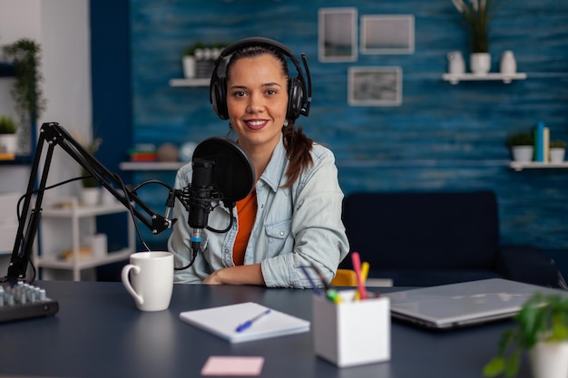 Portrait of woman vlogger wearig headphones and streaming podcast. Social media influencer recording livestream with headphones and microphone in home office. Smiling blogger podcasting.