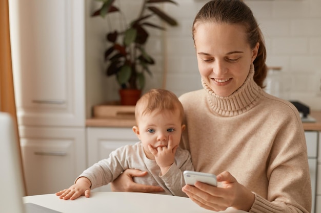 Portrait of woman wearing beige casual style sweater with ponytail hairstyle sitting at table with her infant daughter and using smart phone with positive facial expression.