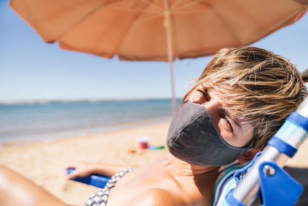 コロナウイルスのパンデミックの真っ只中にあるオレンジ色の傘のすぐ隣のビーチチェアで太陽の下で横たわっている夏休みにフェイスマスクを身に着けている女性の肖像画