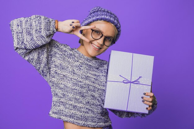 Portrait of a woman wearing knitted hat holding gift box having fun isolated over purple color backg.