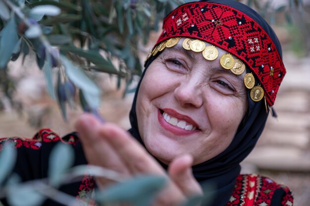 Foto ritratto di una donna che indossa abiti tradizionali palestinesi in un campo di ulivi con un ramo in mano e un sorriso sul viso