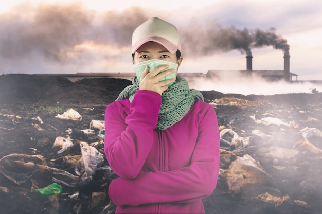 Foto ritratto di una donna che indossa una maschera contro l'inquinamento mentre si trova di fronte a una fabbrica che emette fumo