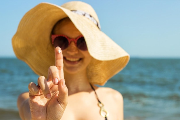 Foto ritratto di una donna che indossa occhiali da sole che mostra la crema sul dito contro il mare e il cielo